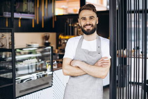 Buchhaltung Gastronomie; Ein Mann in Schürze steht lächelnd, mit Armen verschränkt am Eingang eines Cafés